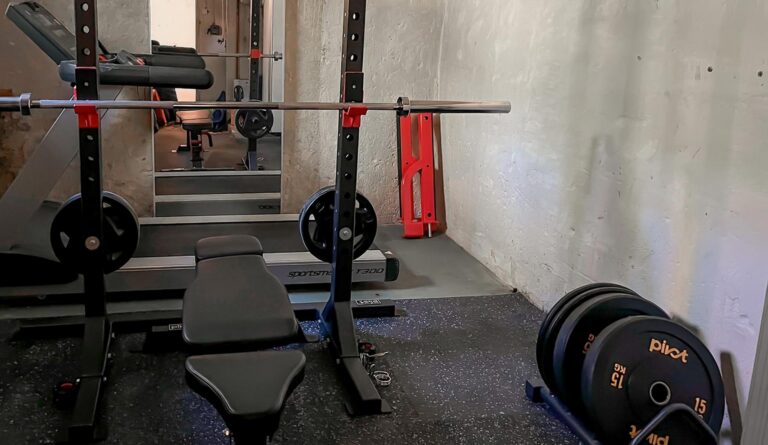 En inzoomad bild av ett hemmagym i källaren med svart gymgolv, skivstångsställning, skivstång, träningsbänk, viktskivor, bumper plates i ställ och ett löpband.