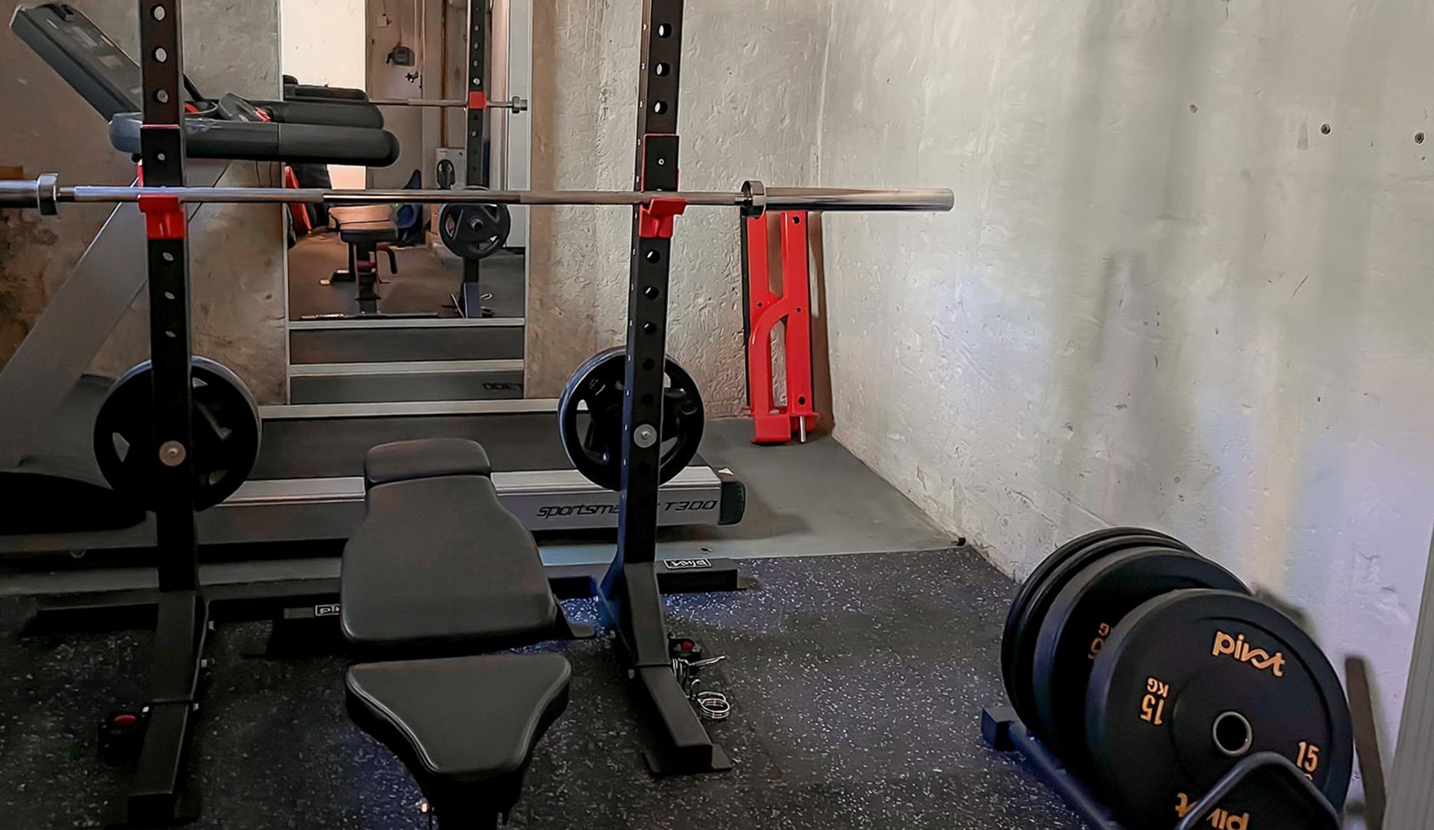 En inzoomad bild av ett hemmagym i källaren med svart gymgolv, skivstångsställning, skivstång, träningsbänk, viktskivor, bumper plates i ställ och ett löpband.